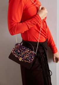 Person som bär en röd långärmad tröja och svart kjol, och håller en svart handväska med en flerfärgad paljettyg och guldkedjeslang.