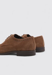 Braune Wildlederschuhe mit glatter Textur, ausgestattet mit klassischem Broguemuster, runder Zehenpartie und brauner Gummisohle.