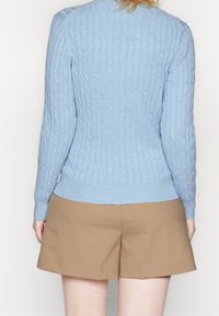 Jasnoniebieski sweter w splot warkoczowy z zaokrąglonym dekoltem i prążkowanymi mankietami, zestawiony z beżowymi szortami o prostym, dopasowanym kroju.