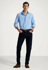 Camisa azul-clara com botões, jeans escuros, cinto de couro castanho e sapatos de camurça castanhos. A camisa possui colarinho e botões na frente.