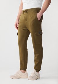 Olivgrüne Jogginghosen aus weichem Stoff, mit einem elastischen Bund und Kordelzug, seitlichen Taschen und einer Utility-Tasche am Bein.
