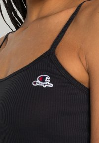 Czarny prążkowany top na ramiączkach z cienkimi paskami, z białym logo "Champion" po lewej stronie. Tekstura wydaje się miękka i dopasowana.
