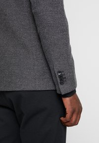 Blazer gris texturizado con un patrón de espiga, que presenta un puño de cuatro botones y una manga interior negra en contraste.