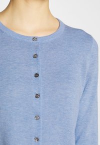 Hellblauer, strickender Cardigan mit rundem Ausschnitt und sechs grauen Knöpfen, die vorne geschlossen sind, getragen am Oberkörper.