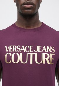Vīrieša tērps: purpursarkans T-krekls ar apļveida kakla izgriezumu, ar lieliem metalizētajiem zelta burtiem "VERSACE JEANS COUTURE" uz krūtīm.