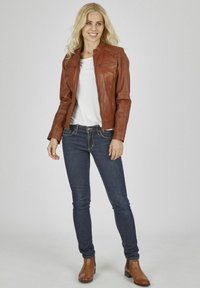 Braune Lederjacke in taillierter Form mit Reißverschluss-Taschen, getragen über einem weißen Oberteil und dunklen Jeans, ergänzt durch braune Stiefeletten.