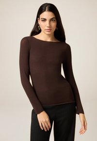 Brown long-sleeve top with a boat neck design, made from a smooth material. Worn with black pinstriped trousers.