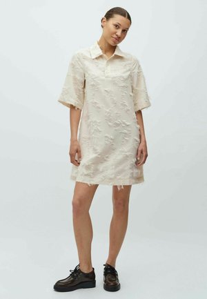 Vestido tipo camisa beige con mangas cortas, tejido texturizado y bordes deshilachados, con cuello de solapa y ajuste suelto. Combinado con zapatos oscuros.