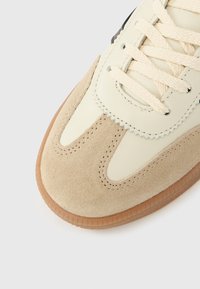 Beige- und cremefarbener Sneaker aus Wildleder und Leder mit Kontrastnähten, runden Schnürsenkeln und einer Gummisohle. Klassisches, flaches Design.
