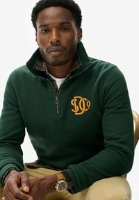 Sudadera de forro polar verde con media cremallera, que presenta un logo bordado en naranja. Debajo, se puede ver un cuello de camisa a cuadros.