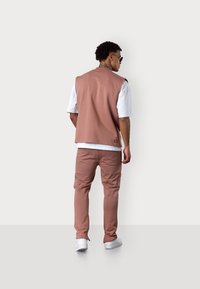 Rosa ärmellose Weste über einem weißen Hemd, kombiniert mit passenden rosa Cargo-Hosen und weißen Sneakers, von hinten vor einem neutralen Hintergrund betrachtet.