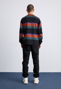 Langærmet sort skjorte med horisontale striber i rust, grøn og blå; parret med sorte sweats og hvide sneakers.