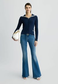 Cardigan blu navy a maglia cable knit con colletto da marinaio bianco e chiusura con bottoni, abbinato a jeans blu svasati e una borsa bianca con accenti dorati.