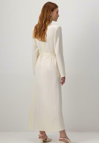 Pikkade punaste juustega naine kannab vööga kreemikat pikkade varrukatega kleiti ja kreemikaid slingback kingi, seistes ühtlase heleda tausta vastas.