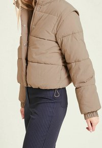 Kort puffjakke i lysebrun nylon med lynlås, trykknapper og ribstrikkede manchetter, kombineret med bukser med nålestriber.