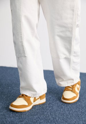 Person iført hvide bukser og brune og hvide sneakers står på et blåt tæppe.
