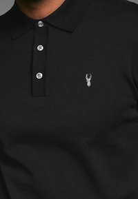 Zwarte poloshirt van textiel met een geribbelde kraag, drie knopenlijst en een klein geborduurd hertlogo op de borst.