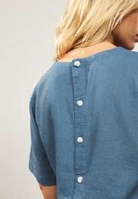 Blue linen shirt with short sleeves, round neckline, and a button-down back featuring five white buttons along the center.