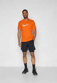 Oranje Nike T-shirt met een meerkleurig logo, gecombineerd met zwarte shorts. Zwarte sportschoenen en sokken maken de outfit compleet.
