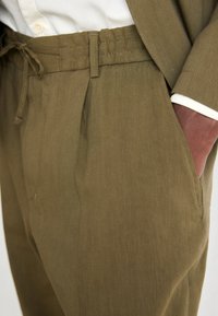 Olive green linen trousers with a drawstring waistband, pleats, side pockets, and a smooth texture. White shirt cuffs are visible.