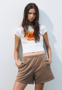 T-shirt bianca corta con una stampa di girasole sulla parte anteriore; abbinata a pantaloni corti in maglia marroni con tasche laterali e un elastico in vita.