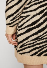 Pull beige à rayures zébrées noires. Poignets et ourlet côtelés, texture douce, et détail fendu sur le côté. Main touchant délicatement le tissu.