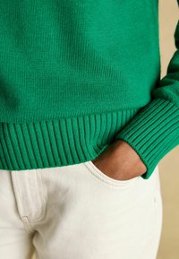 Grüner gestrickter Pullover mit gerippten Bündchen und Saum, kombiniert mit cremefarbener Hose mit Seitentasche. Fokus auf Textur und Farbe.