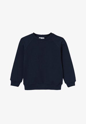 Chłopiec uśmiechający się, ubrany w granatową bluzę, jasnoniebieskie dżinsy i białe trampki, stojący na tle jednolitego jasnoszarego tła.