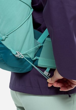 Person, die an der Kordel des türkisfarbenen Rucksacks zieht, der über einer lila Jacke und hellgrünen Hosen getragen wird; der Fokus liegt auf der Hand und den Rucksackriemen.