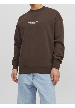 La personne porte un sweat-shirt marron avec le texte « originals studio » sur la poitrine et un jean bleu clair sur un fond gris uni.