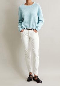 Ljust blå stickad tröja med texturfinish, vita slim-fit jeans och svarta vävda sandaler; modellen har händerna i fickorna, neutral bakgrund.