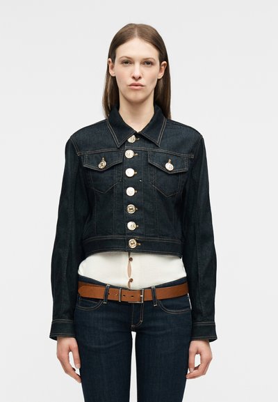 Γυναίκα που φοράει σκούρο τζιν μπουφάν με μεγάλα διακοσμητικά χρυσά κουμπιά, συνδυασμένο με σκούρο τζιν, καφέ ζώνη και λευκό τοπ από κάτω.