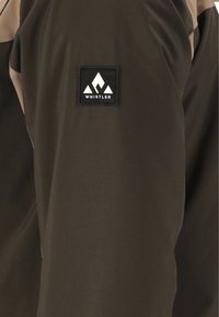 Dettaglio di una giacca marrone scuro con una toppa quadrata nera che include una grafica di una montagna bianca. Il tessuto è liscio con texture minima.