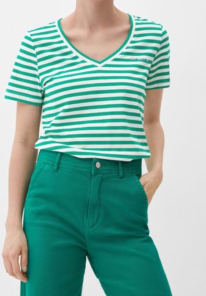Persoon draagt een groen-wit gestreept V-hals shirt, ingestopt in een high-waisted effen groene broek, met één hand in de zak.