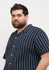 Camisa de manga corta en azul marino con rayas verticales blancas, con cuello y cierre de botones. Tejido texturizado con un corte holgado.