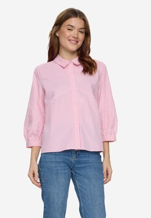 Frau trägt eine rosa-weiß gestreifte Langarmbluse mit Kragen und blaue Jeans, steht vor einem schlichten Hintergrund.