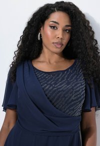 Dunkelblaues Kleid mit einem strukturierten, schimmernden Oberteil im Wellenmuster. Kurze, schwingende Ärmel und ein drapierter Ausschnitt verleihen Weichheit.