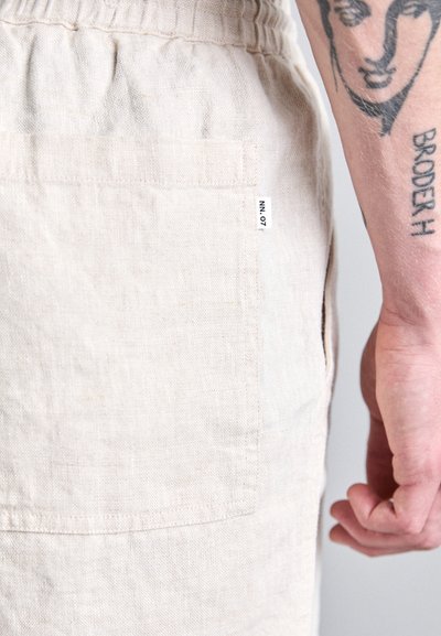 Shorts en lin beige avec une poche arrière et une étiquette portant "NN.07." Le tissu a une apparence texturée, des coutures visibles et une coupe douce.