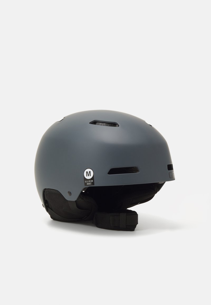 Casco grigio opaco con fessure di ventilazione, cinturino regolabile e etichetta di taglia (M, 55-59 cm). Superficie liscia e design arrotondato.