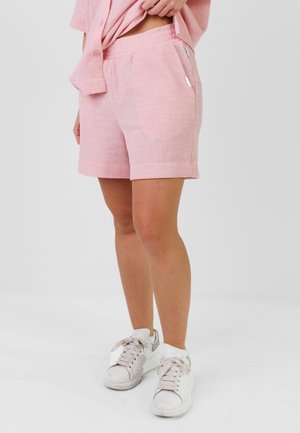 Personne portant un short texturé rose et un haut assorti, associé à des baskets blanches, debout devant un fond clair uni.