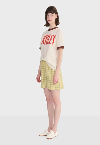 Mladá žena stojaca, oblečená v krémovom tričku s červeným nápisom a lemom, žltou károvanou sukňou a bielymi šnurovacími topánkami na jednofarebnom pozadí.