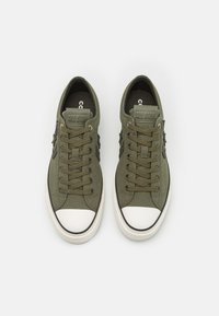 Sneakers in tela verde oliva con punte in gomma bianca, lacci rotondi e dettagli neri. Presentano una patch del logo sulla linguetta.