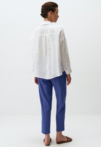 Witte linnen shirt met een ontspannen pasvorm en rugpand, gecombineerd met slim-fit blauwe broek. Model draagt bruine sandalen en staat op een neutrale achtergrond.