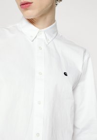 Chemise blanche en coton à boutons avec un col classique, boutons visibles et un petit logo noir sur la poitrine.