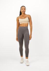 Sujeción deportiva beige con banda del logo, combinada con leggings gris ajustados y de cintura alta, todo hecho de una tela elástica, y llevada con zapatillas de color claro.