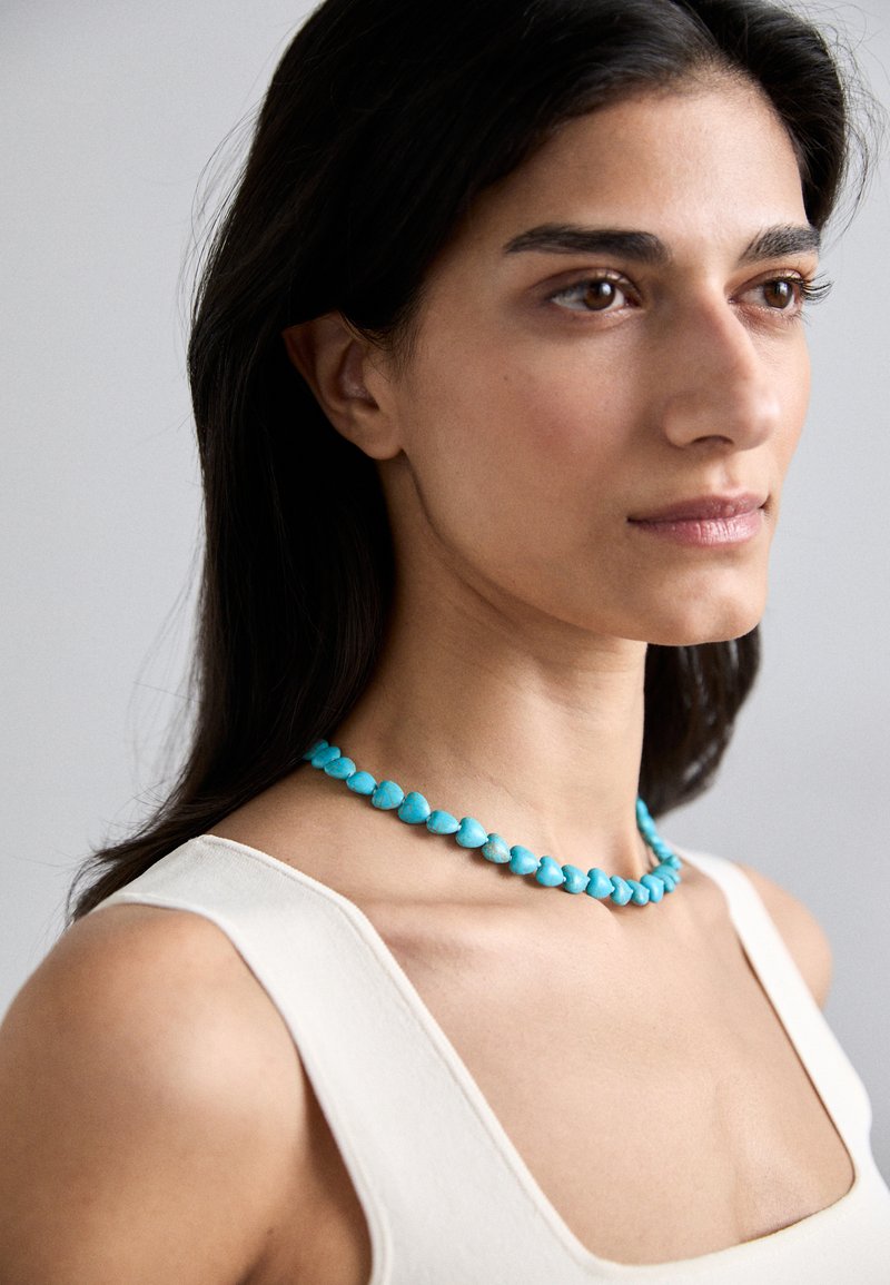 Τουρνουάζ κολιέ με καρδιοειδή χάντρες. Λείο άγγι�γμα, ζωντανό χρώμα και ομοιόμορφα κατανεμημένος σχεδιασμός που φοριέται πάνω από ένα ελαφρύ, αμάνικο τοπ.