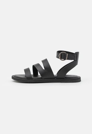 Sort flad sandal med tre læderremme hen over foden og en ankelrem med en sølvspænde på en hvid baggrund.