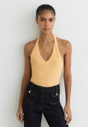 Kobieta z warkoczem, ubrana w bezrękawnikowy brzoskwiniowy top typu halter i ciemnoniebieskie, wysokie spodnie jeansowe z przednimi kieszeniami, stojąca przy białej ścianie.