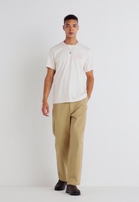 Light beige t-shirt with short sleeves and a small pink graphic, paired with khaki wide-leg pants and dark brown shoes. Minimalist design.