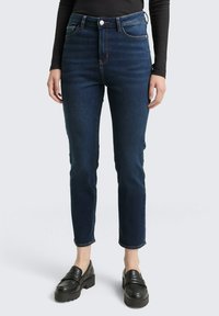 Naisso, kes kannab tumedatest sinist õhukest istuvat teksapükse, musta pikade varrukate topi ja musti platvormide loofereid, seisab lihtsa tausta vastas.